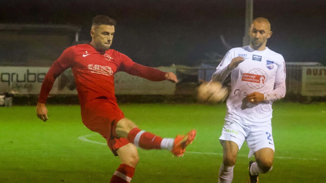 Zwei Fußballspieler auf einem Feld. Einer in Rot tritt den Ball, der andere in Weiß verteidigt.