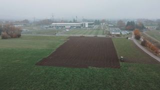Eine Draufsicht eines nebligen Feldes mit einem Traktor und einem Baufahrzeug. Im Hintergrund sind Gebäude und geparkte Autos zu sehen.