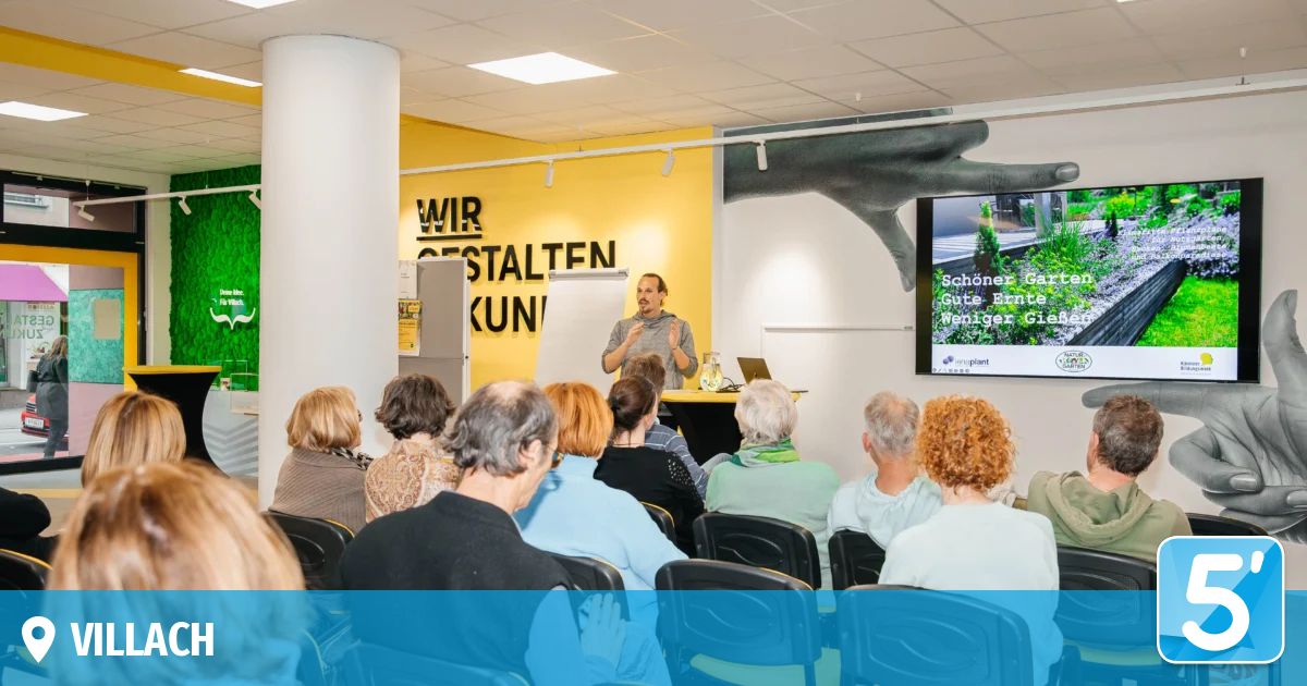 Ein Mann spricht zu einem sitzenden Publikum in einem Konferenzraum. Die Wände sind gelb mit einer großen Grafik. Ein Monitor zeigt eine Nachricht an.