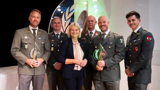 Eine Gruppe von fünf Personen in Militäruniformen steht zusammen und lächelt. Sie halten Trophäen. Eine Frau in der Mitte trägt einen Anzug und einen Schal. Sie befinden sich wahrscheinlich bei einer Preisverleihung. Hinter ihnen ist eine grüne Wand mit einer Lampe.