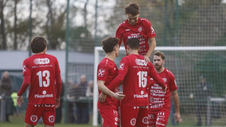 Vier Fußballspieler in roten Trikots feiern auf einem Feld. Ein Spieler wird von anderen hochgehoben. Sie tragen die Nummern 15, X und 2. Ein Tor steht hinter ihnen.