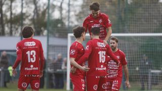 Vier Fußballspieler in roten Trikots feiern auf einem Feld. Ein Spieler wird von anderen hochgehoben. Sie tragen die Nummern 15, X und 2. Ein Tor steht hinter ihnen.