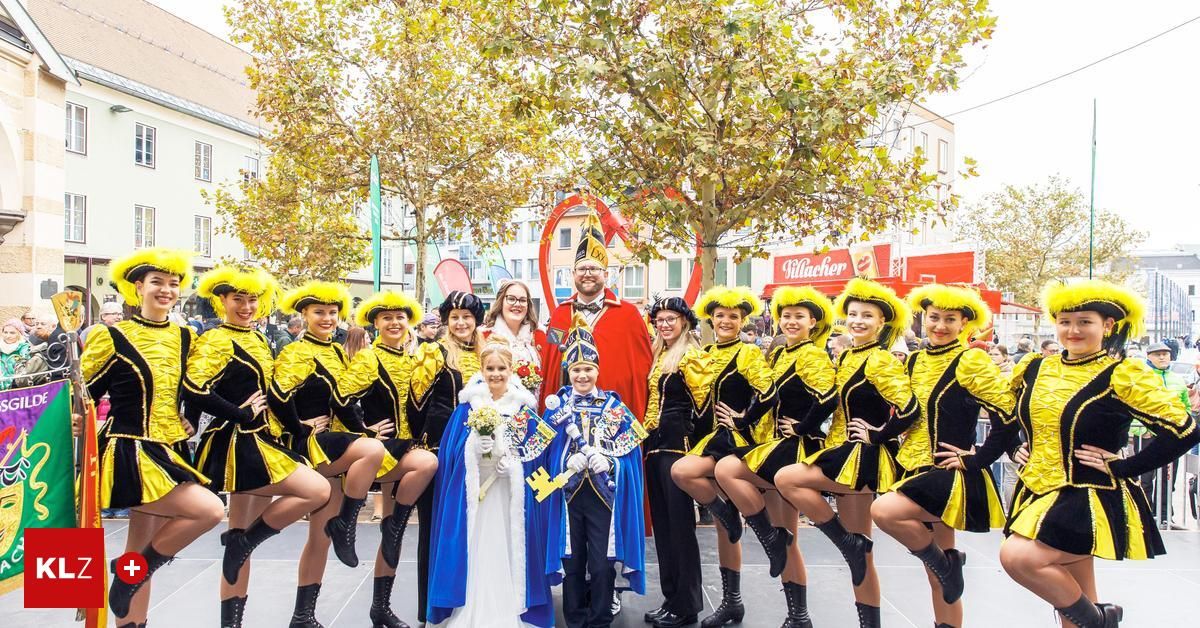 Eine Gruppe von Menschen in Kostümen posiert für ein Foto auf einer Bühne, darunter eine Braut, ein Junge und andere. Dahinter steht ein Baum, ein Gebäude und ein Schild mit der Aufschrift "Villacher".