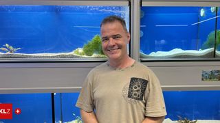 Ein Mann steht lächelnd vor zwei Aquarien mit blauem Wasser und grünen Pflanzen. Er trägt ein beiges T-Shirt mit schwarzem Schlangengrafik.