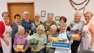 Eine Gruppe älterer Frauen lächelt in einem Raum und hält Boxen mit Noe-Plätzchen. Eine Frau hält einen Blumenstrauß. Dahinter hängen zwei gerahmte Bilder an einer weißen Wand.