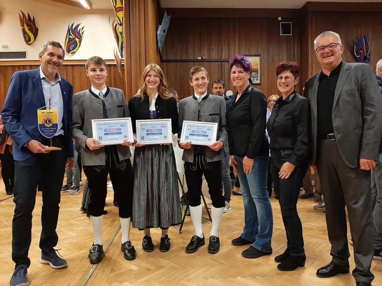 Eine Gruppe von Menschen steht in einem Raum und hält Urkunden. Drei Personen halten große Urkunden, während andere im Hintergrund lächeln. Der Raum hat Holzwände und -böden.