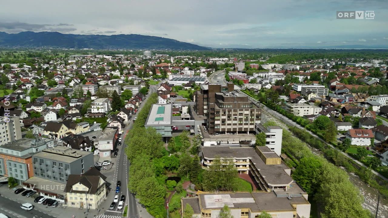 Luftaufnahme einer Stadt mit vielen Gebäuden, Bäumen und Bergen in der Ferne. Eine breite Straße mit Autos und einem Rasenbereich.