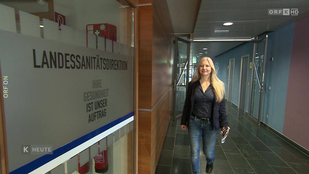 Eine Frau geht einen Flur entlang mit einem Schild, auf dem steht 'Ihre Gesundheit ist unsere Aufgabe.' Sie trägt eine Mappe und scheint auf dem Weg zu einem Büro zu sein.