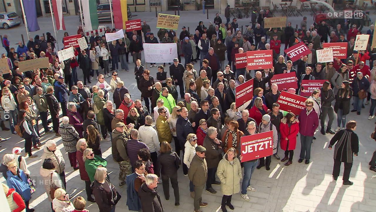 Eine große Menschenmenge versammelt sich auf einem Freigelände, wahrscheinlich bei einer Demonstration. Sie halten rote Schilder mit weißem Text in den Händen. Einige der Schilder tragen die Aufschriften "JETZT!" und "Verdient mehr!". Die Menschen tragen verschiedene Kleidungsstücke, darunter Hüte, Brillen und Sneaker. In der Ferne befinden sich Gebäude und geparkte Fahrräder.