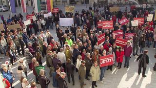 Eine große Menschenmenge versammelt sich auf einem Freigelände, wahrscheinlich bei einer Demonstration. Sie halten rote Schilder mit weißem Text in den Händen. Einige der Schilder tragen die Aufschriften "JETZT!" und "Verdient mehr!". Die Menschen tragen verschiedene Kleidungsstücke, darunter Hüte, Brillen und Sneaker. In der Ferne befinden sich Gebäude und geparkte Fahrräder.