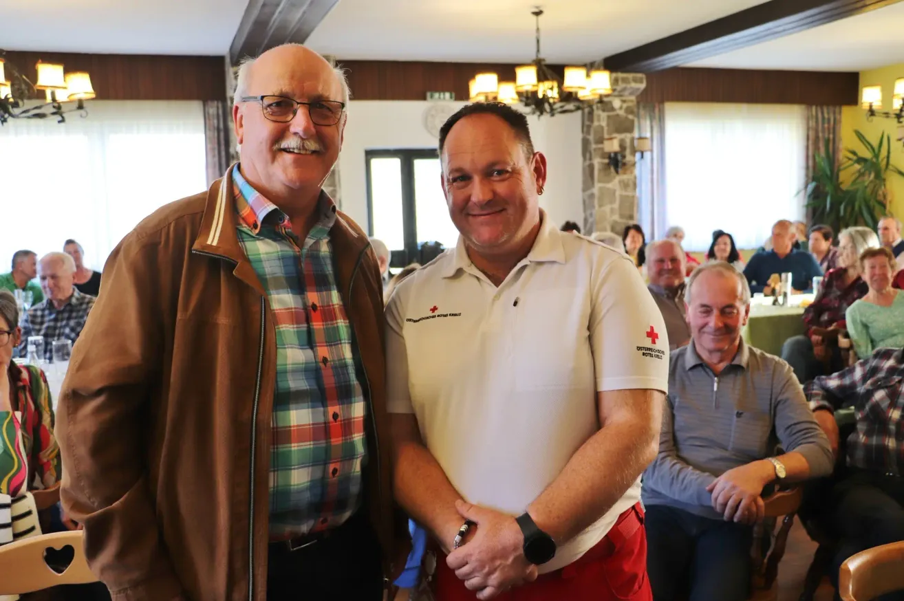 Zwei Männer stehen zusammen und lächeln für ein Foto. Einer trägt eine braune Jacke und ein kariertes Hemd, der andere ein weißes Poloshirt mit rotem Kreuzemblem. Hinter ihnen sitzt ein Publikum und beobachtet.