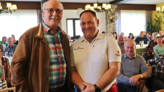 Zwei Männer stehen zusammen und lächeln für ein Foto. Einer trägt eine braune Jacke und ein kariertes Hemd, der andere ein weißes Poloshirt mit rotem Kreuzemblem. Hinter ihnen sitzt ein Publikum und beobachtet.