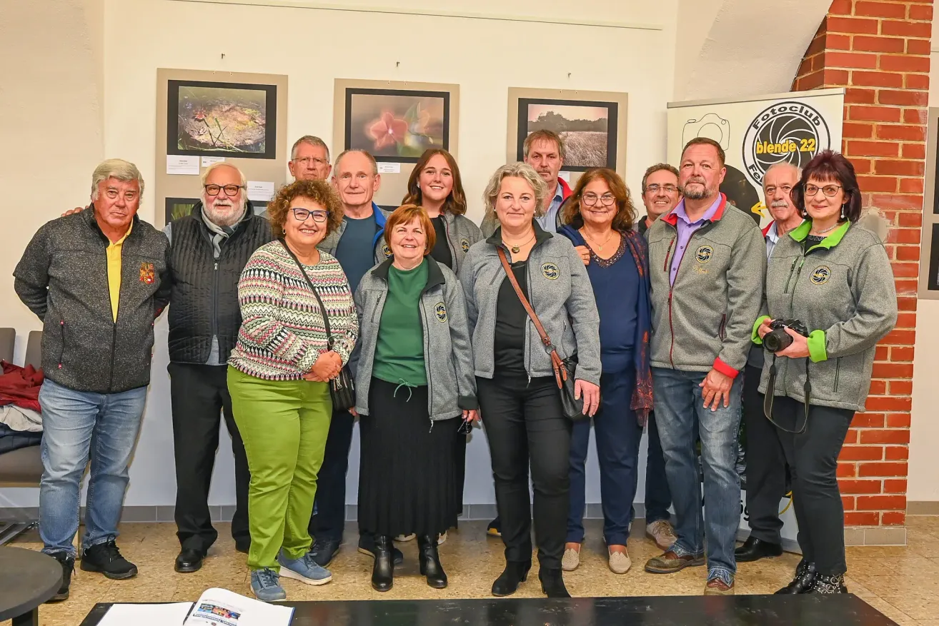 Eine Gruppe von Menschen posiert für ein Foto vor einer Wand mit verschiedenen gerahmten Bildern. Sie lächeln und scheinen glücklich zu sein.