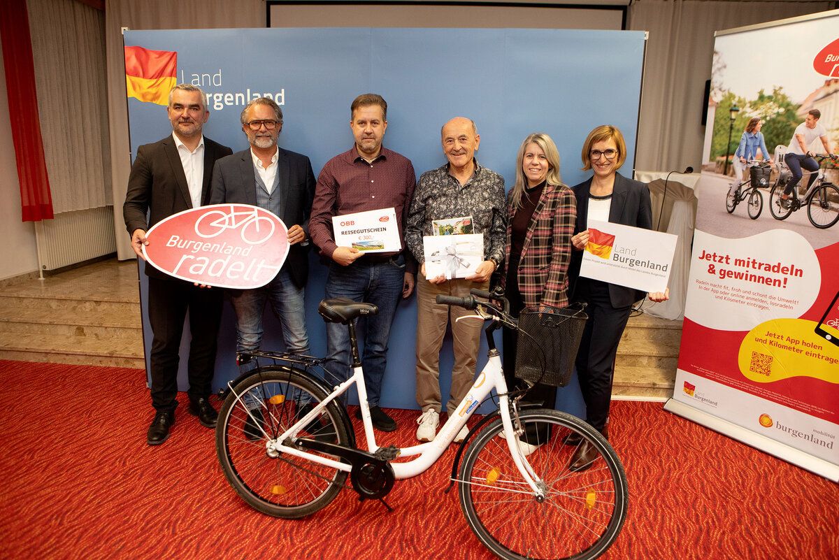 Eine Gruppe von Menschen steht um ein Fahrrad herum, einer Mann hält eine Geschenkbox und ein anderer eine Flagge. Sie alle lächeln und posieren für ein Foto.