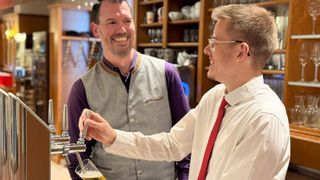 Zwei lächelnde Männer in einer Bar, einer hält einen Bierhahn, der andere mit einem Glas Bier. Sie sind von Regalen mit Gläsern und Tassen umgeben.