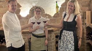 Drei Frauen halten Weingläser in der Hand, lächeln in einem Raum mit Backsteinwänden, Kamin und Weinflaschen.