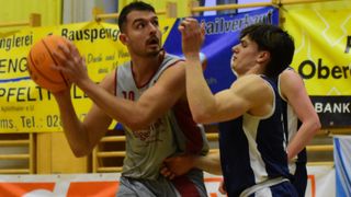 Zwei Basketballspieler spielen in einer Halle. Ein Spieler in einem grauen Trikot hält den Ball, während er von einem anderen Spieler in einem blauen Trikot bewacht wird. Dahinter ist teilweise ein gelbes Banner zu sehen.