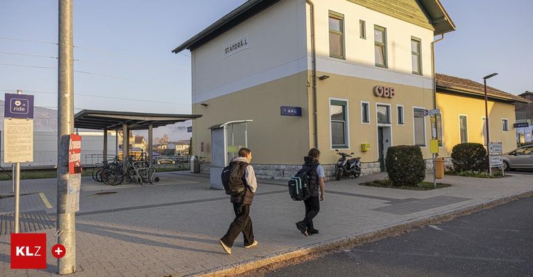 Zwei Personen mit Rucksäcken gehen auf dem Bürgersteig vor einem Bahnhof. Ein Roller ist draußen geparkt.