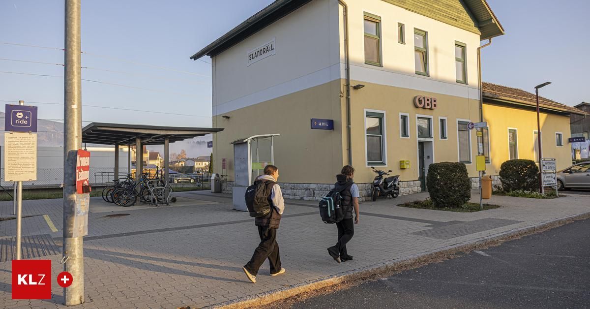 Zwei Personen mit Rucksäcken gehen auf dem Bürgersteig vor einem Bahnhof. Ein Roller ist draußen geparkt.