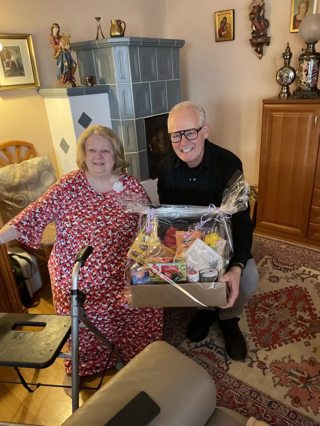 Eine ältere Frau und ein Mann in einem schwarzen Shirt halten eine Geschenkbox mit Plastikfolie, stehend in einem Wohnzimmer mit einem Kamin.