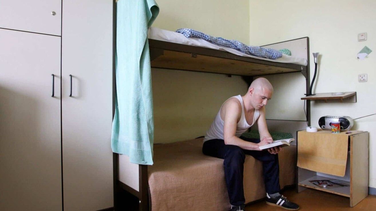 Ein Mann sitzt auf einem unteren Etagenbett in einem Zimmer und liest ein Buch. Ein Handtuch hängt an der Seite des Bettes.