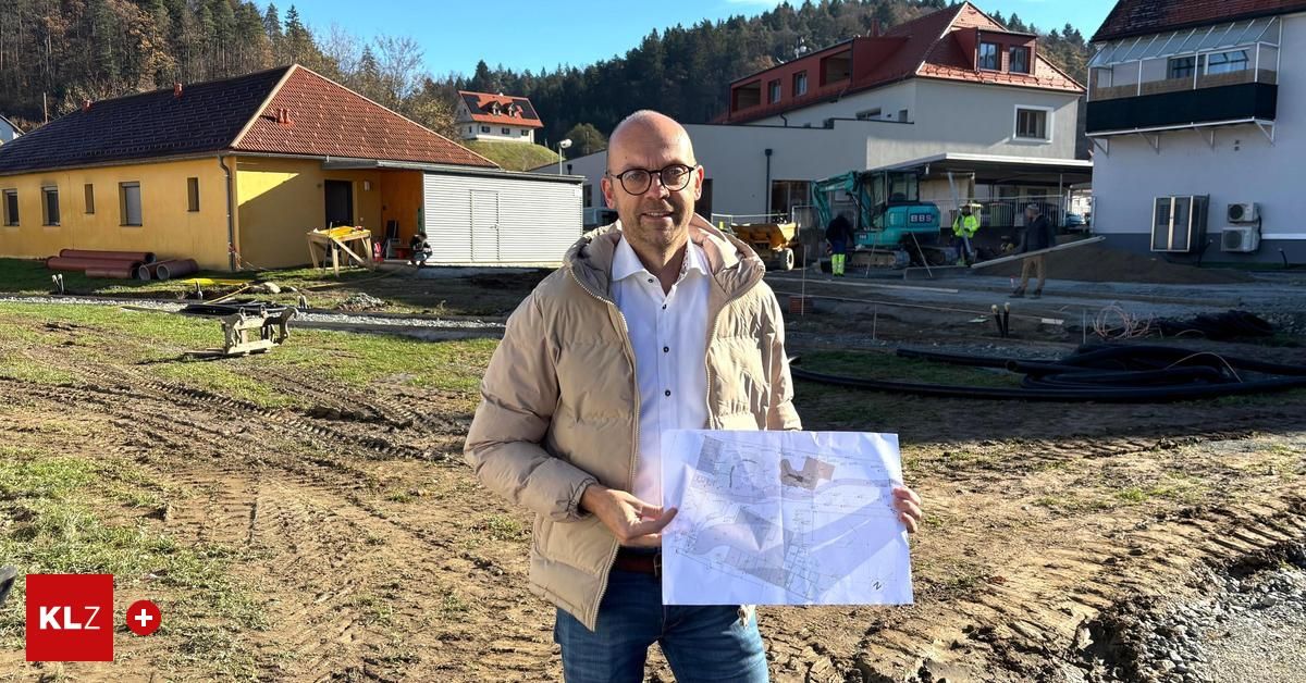 Ein Mann in einer beigen Jacke steht auf einer Baustelle und hält einen Bauplan in der Hand. Hinter ihm sind Häuser, ein Baufahrzeug und Arbeiter zu sehen. Die Baustelle ist von Gras und Erde umgeben.