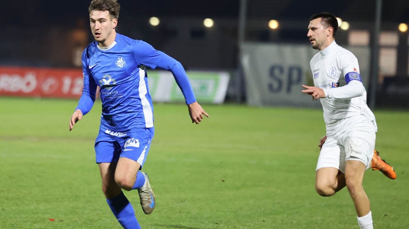 Ein Fußballspieler in blauer Uniform läuft auf dem Feld, mit einem anderen Spieler in Weiß hinter ihm. Beide tragen Turnschuhe. Der Hintergrund zeigt ein Banner mit den Buchstaben SP.
