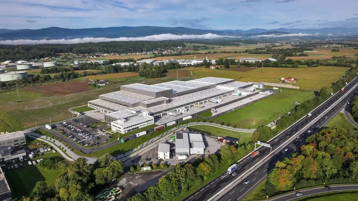 Luftaufnahme einer großen Industrieanlage mit mehreren Gebäuden, einem Parkplatz und einer nahe gelegenen Autobahn. Das Gebiet ist von Bäumen und Gras umgeben, mit Bergen in der Ferne.