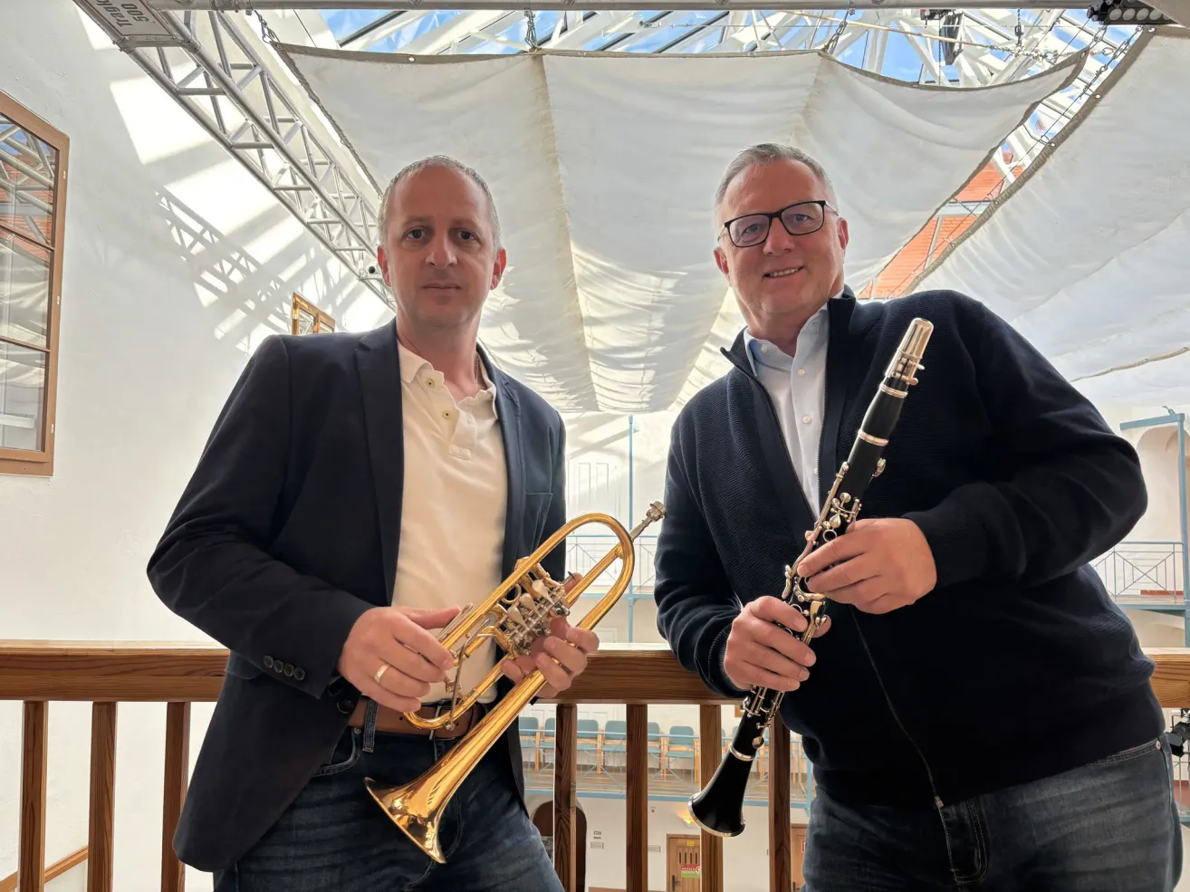 Zwei Männer in Anzügen stehen nebeneinander, einer hält eine Trompete und der andere eine Klarinette und lächeln für ein Foto in einem Musikstudio mit weißer Decke.