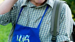 Ein lächelnder Mann mit einer blauen Schürze, auf der 'Luis aus Schlittl' steht, hält eine Karte in der Hand und telefoniert. Er steht auf einer Rasenfläche.