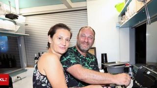 Ein Mann und eine Frau lächeln in einer Küche. Der Mann hält einen Topf auf einem Herd, während die Frau neben ihm steht. Hinter ihnen befindet sich ein Regal mit Flaschen und Behältern.