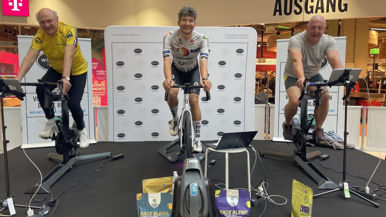 Ein Mann fährt ein Fahrrad in einem Geschäft mit einem Schild hinter ihm, das 'Gentlemen's Choice für Soundgewinner' sagt. Er trägt ein Radtrikot und hält das Lenkrad. Es gibt einen Laptop, Taschen und eine Flasche auf der Bühne.