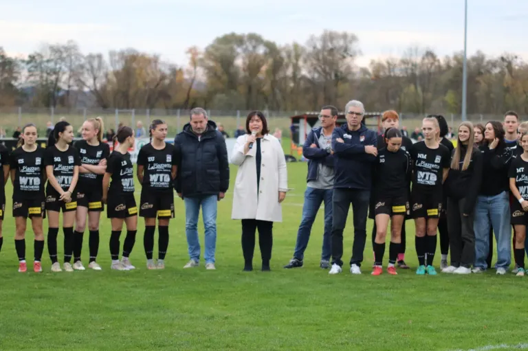 Eine Gruppe von Fußballspielern, darunter eine Frau mit einem Mikrofon, steht zusammen auf einem Rasenfeld. Hinter ihnen stehen mehrere Männer mit verschränkten Armen. Im Hintergrund befinden sich Bäume und ein Zaun.