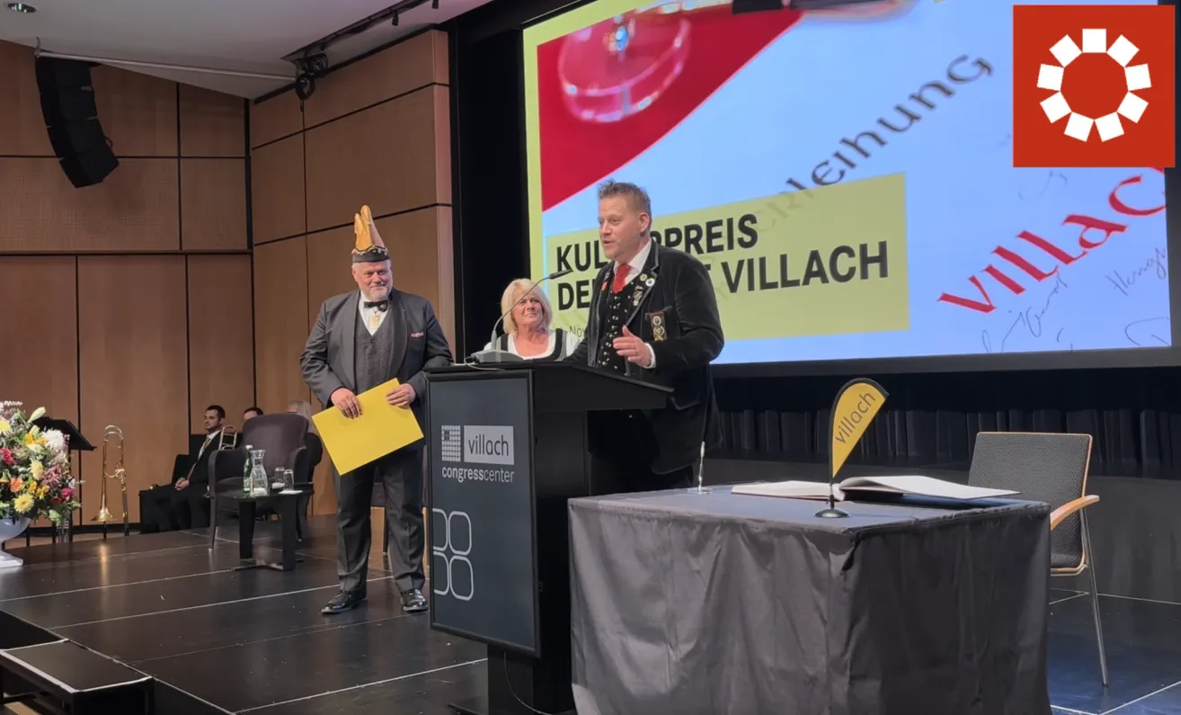 Ein Mann steht an einem Podium in einem Konferenzraum und hält eine gelbe Mappe. Hinter ihm sitzt eine Frau an einem Schreibtisch. Ein Mann in einem Hut steht neben ihm. Eine Projektionsleinwand zeigt die Worte 'KULTUR PREIS VILLACH'.