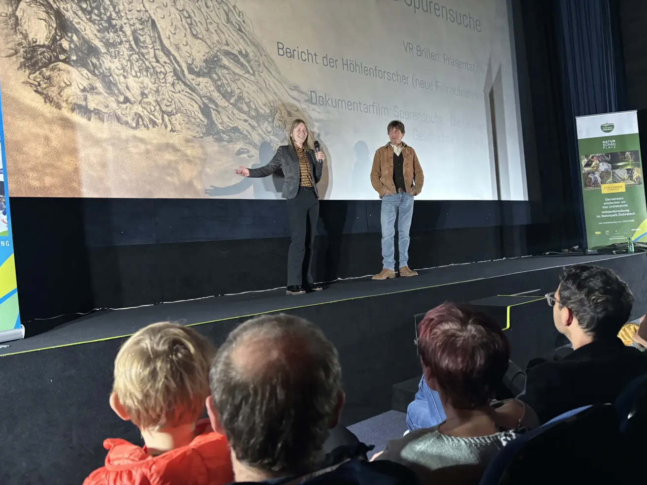 Zwei Personen stehen auf der Bühne vor einer Projektionsleinwand mit einer Berglandschaft, eine Frau hält ein Mikrofon und lächelt, während ein Mann neben ihr steht. Ein Publikum sitzt vor ihnen.