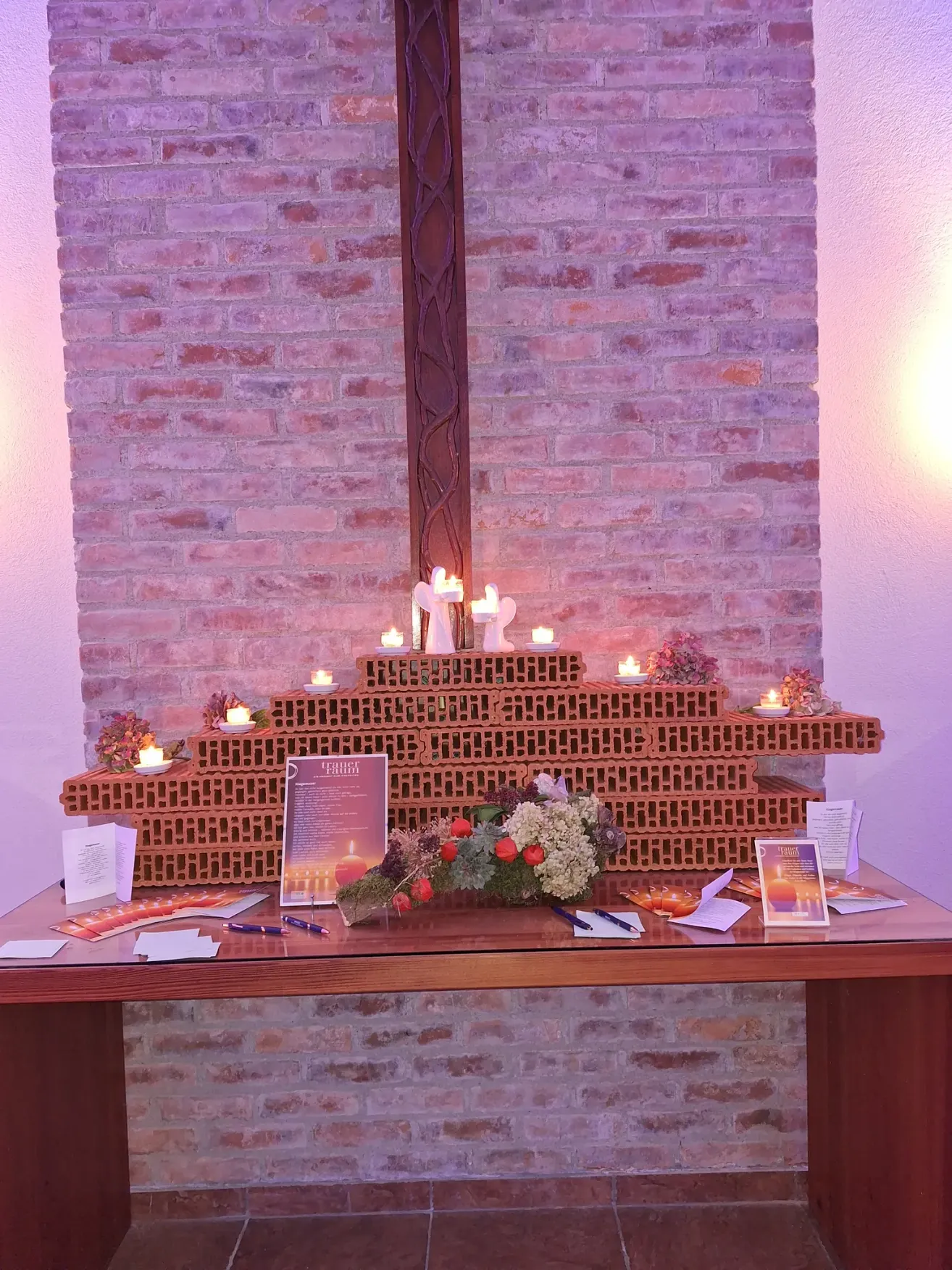 A brick altar with several candles and flowers, a program, and pens on a desk in a pink-lit room.