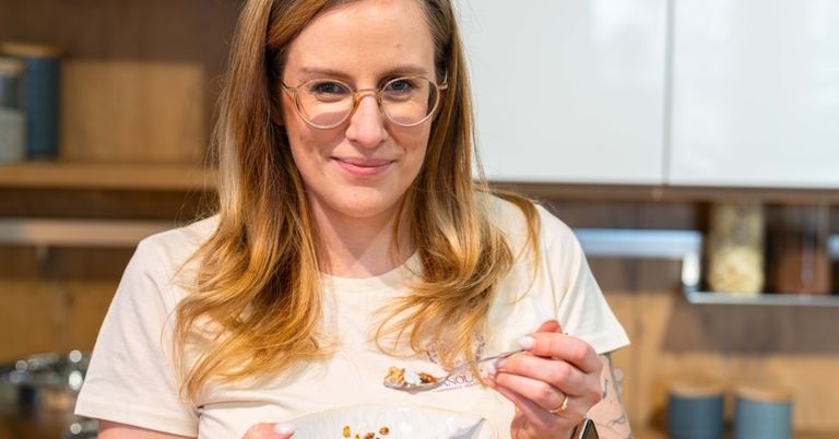 Eine Frau mit Brille lächelt, während sie einen Löffel mit Essen hält, ein weißes T-Shirt mit einem Design trägt und einen Ring am Finger hat.