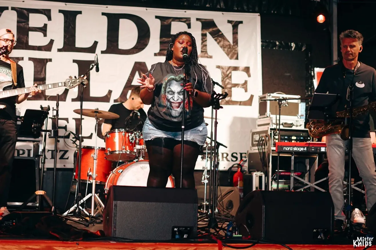 Eine Frau singt auf der Bühne mit einem Mikrofon. Hinter ihr spielt ein Schlagzeuger ein Schlagzeug. Ein Banner mit dem Wort 'GOLDEN' hängt an der Wand.