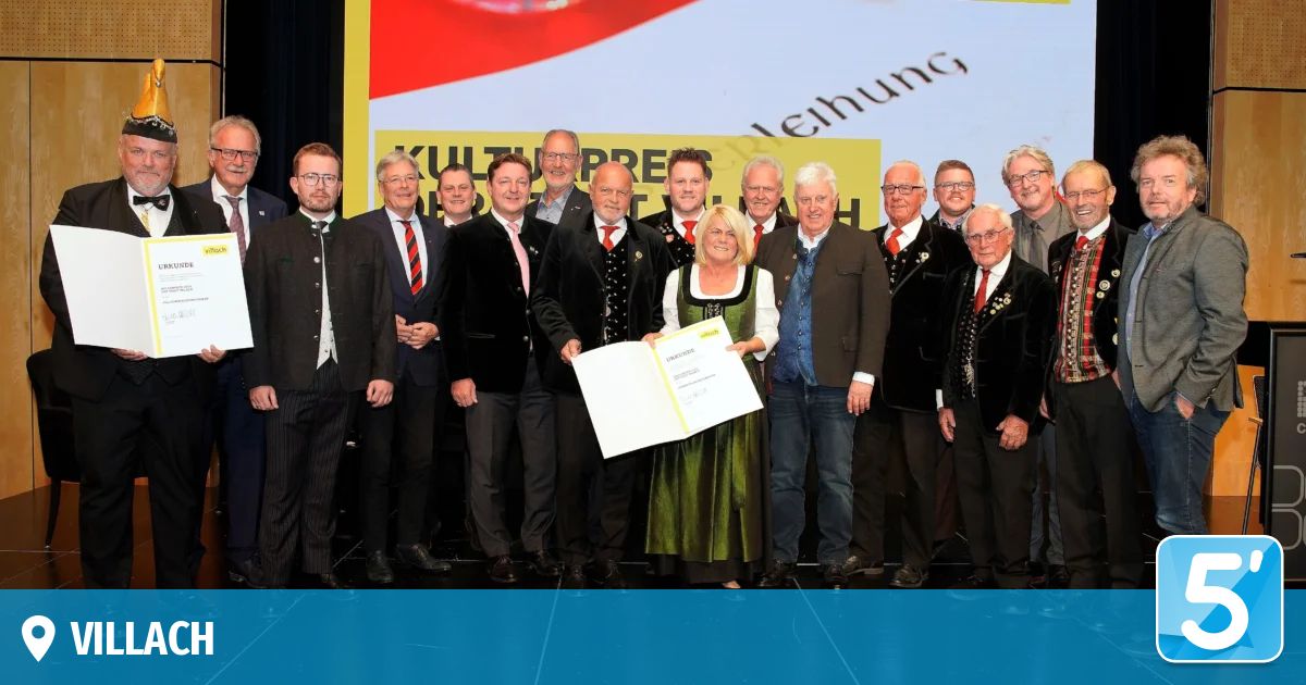 Eine Gruppe formell gekleideter Männer und Frauen steht auf einer Bühne, wobei eine Frau ein Zertifikat in der Hand hält. Sie scheinen für ein Foto zu posieren. Hinter ihnen ist ein Hintergrund mit deutschem Text.