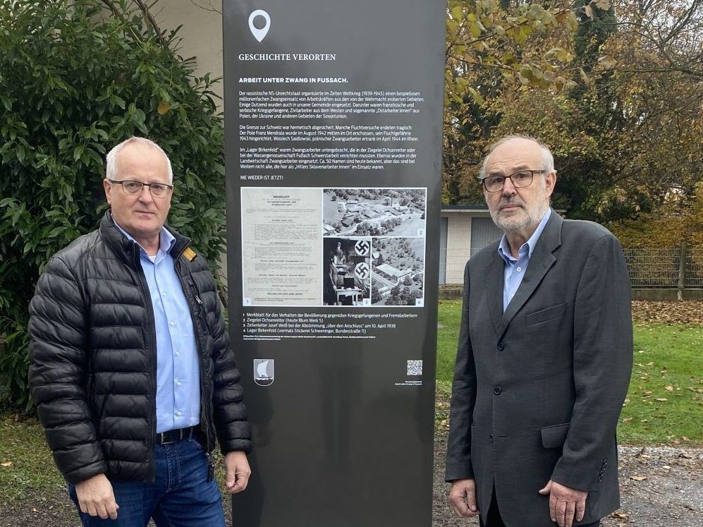 Two men stand in front of a monument. The sign reads 'History Unearthed' and 'Work Under Compulsion in Füssen'. The man on the left wears a black jacket and glasses. The man on the right wears a dark suit.
