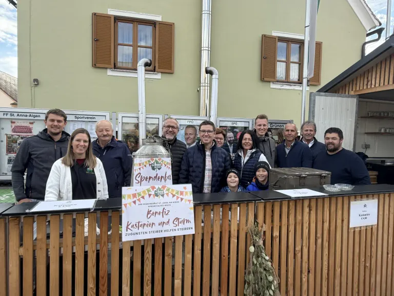 Eine Gruppe von Menschen steht vor einem Gebäude und lächelt für ein Foto. Im Vordergrund befindet sich ein Holzzaun mit einem Schild, auf dem Kastanien und Sturm steht.