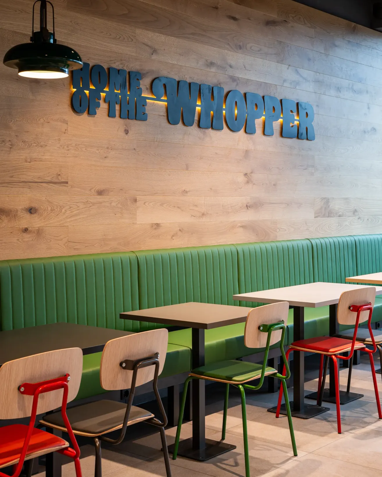 Ein leeres Restaurant mit Holzwänden hat grüne Sitzbänke, Holztische und farbenfrohe Stühle. Die Wand zeigt die Worte 'Home of the Whopper'. Eine Lampe ist an der Decke beleuchtet.
