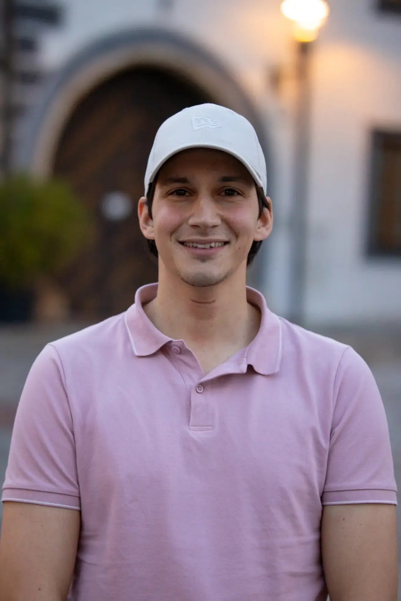 Ein Mann in einem rosa Poloshirt und einer weißen Baseballkappe lächelt im Freien.