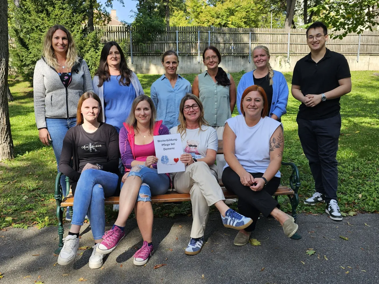 Eine Gruppe von Personen sitzt auf einer Parkbank und hält ein Schild mit der Aufschrift 'Weiterbildung - Pflege bei Demenz.' Sie sind lässig gekleidet. Dahinter grenzt ein Holzzaun ein Grasfeld mit Bäumen ein.