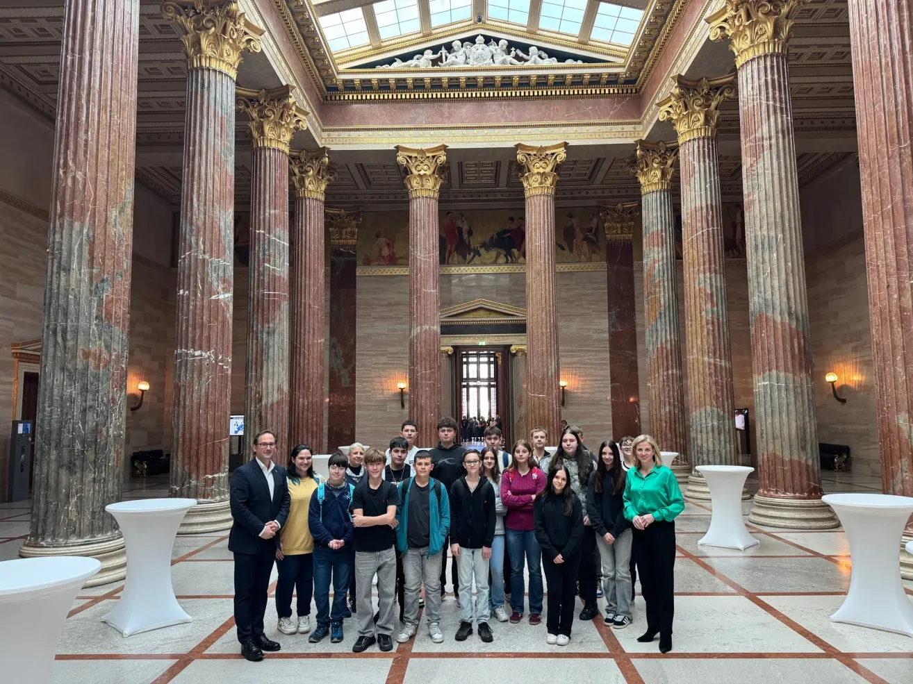 Eine Gruppe von Schülern und Lehrern steht in einem großen, reich verzierten Saal mit hohen Marmorsäulen und einer Glasdecke. Einige Schüler tragen Freizeitkleidung, und eine Frau in Grün hält ein Mikrofon.