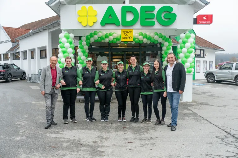 Eine Gruppe von Menschen in passenden Uniformen steht vor einem Geschäft mit einem grün-weißen Banner darüber. Das Schild heißt ADEG. Das Geschäft hat grüne und weiße Luftballons davor.