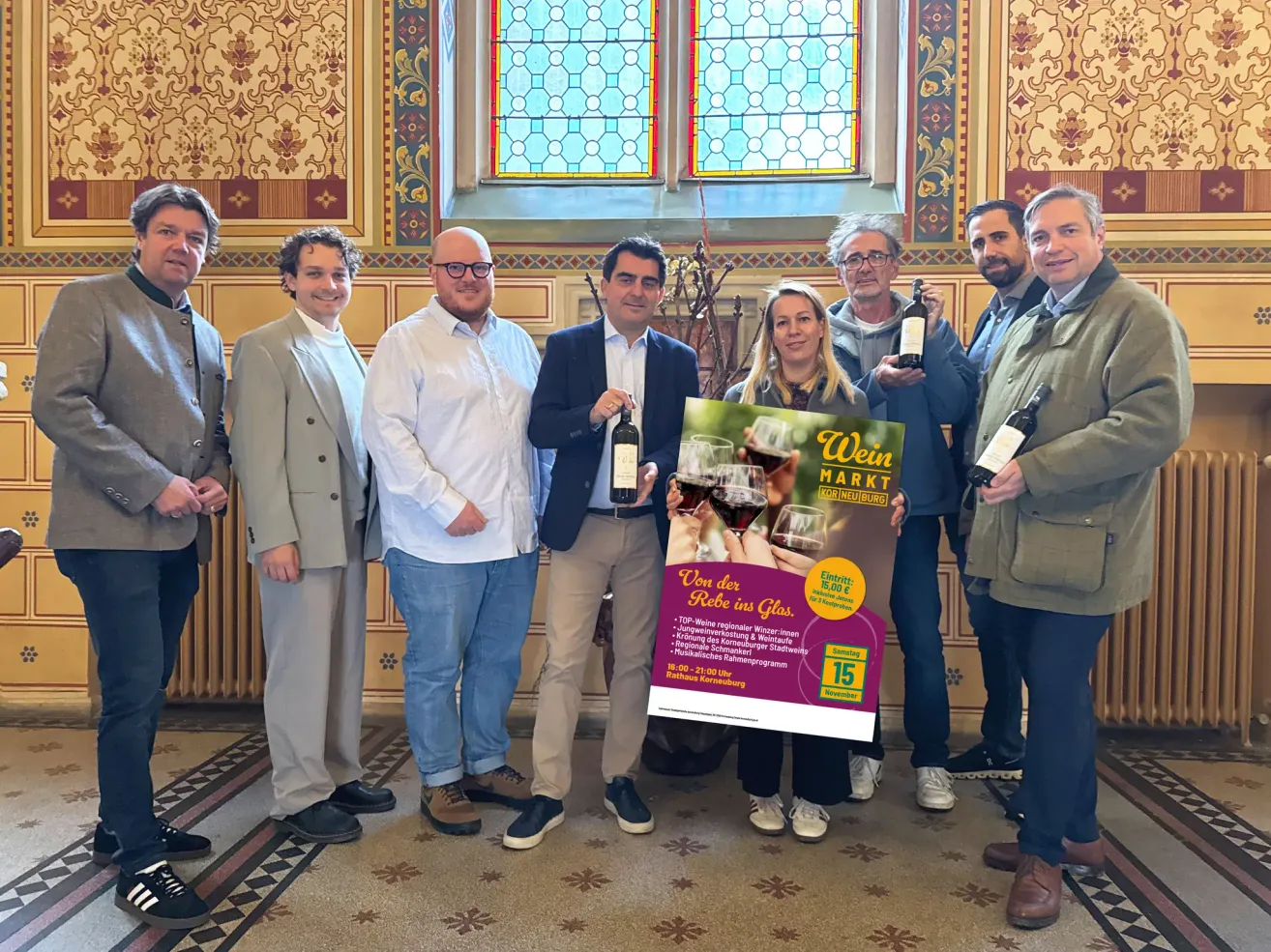 Eine Gruppe von Menschen steht vor einem farbenfrohen Hintergrund, hält Weinflaschen und lächelt für ein Foto. Die Frau in der Mitte hält ein Plakat.