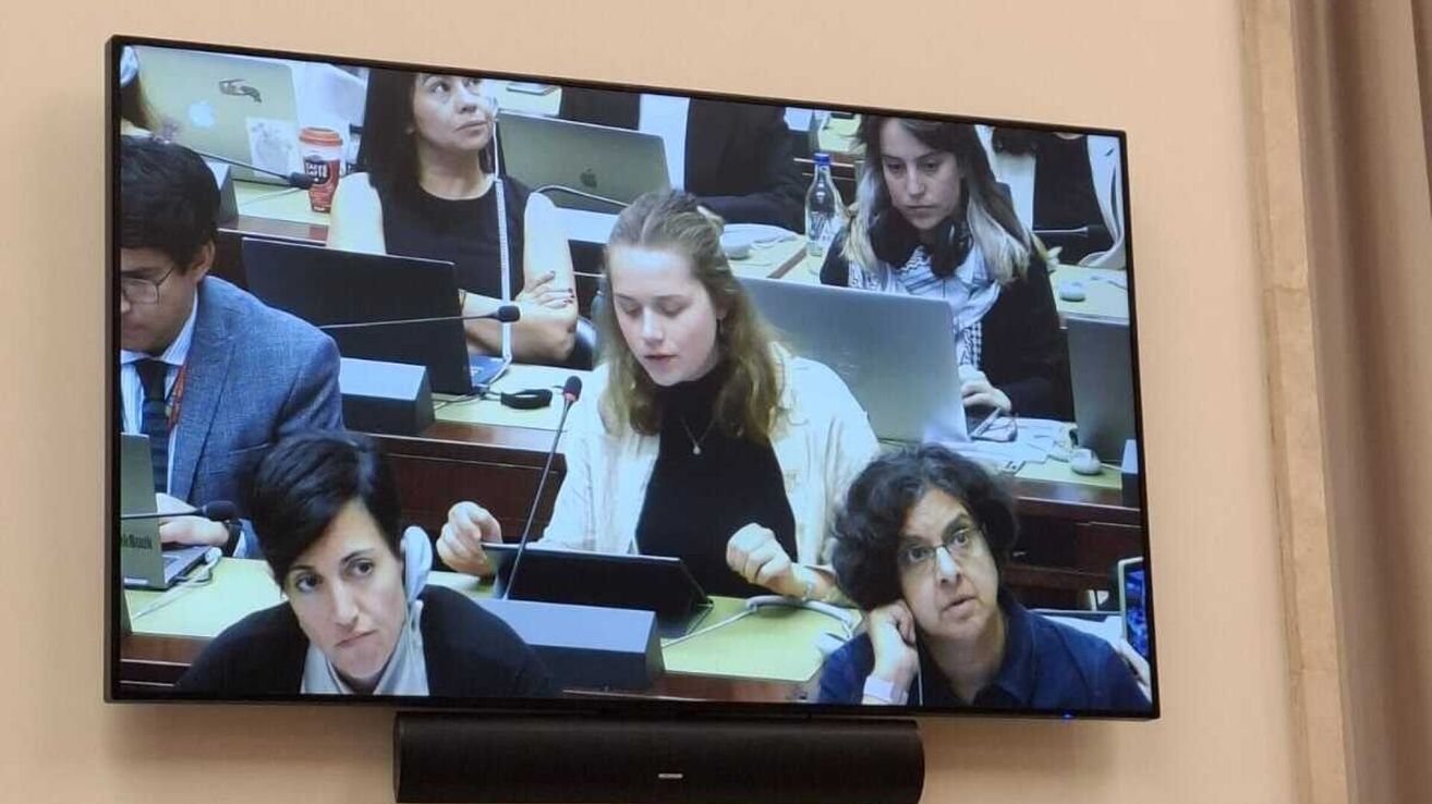 Ein Konferenzraum mit mehreren Frauen an Schreibtischen, eine Frau im Vordergrund spricht an einem Podium. Andere schauen auf Laptops. Ein Mikrofon und ein Laptop sind auf dem Tisch vor ihnen.