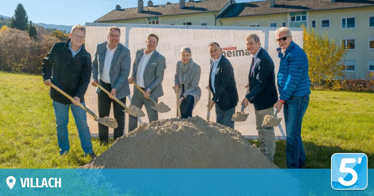 Eine Gruppe von Personen mit Schaufeln steht auf einem Rasen, wahrscheinlich bei einer Grundsteinlegungszeremonie, mit einem Haufen Sand vor ihnen. Dahinter befindet sich ein Banner mit Text und Logos.