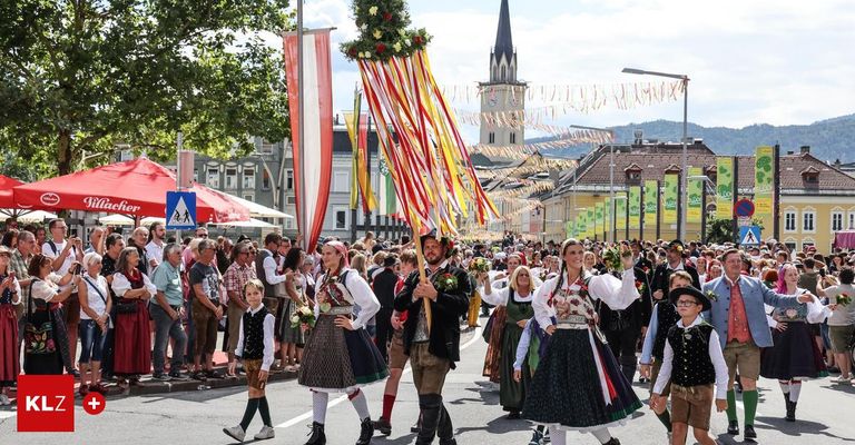 Ein festlicher Umzug mit Menschen in traditionellen Kostümen, die mit Blumen und Bannern durch die Straße ziehen. Im Hintergrund befinden sich Gebäude und ein Turm.